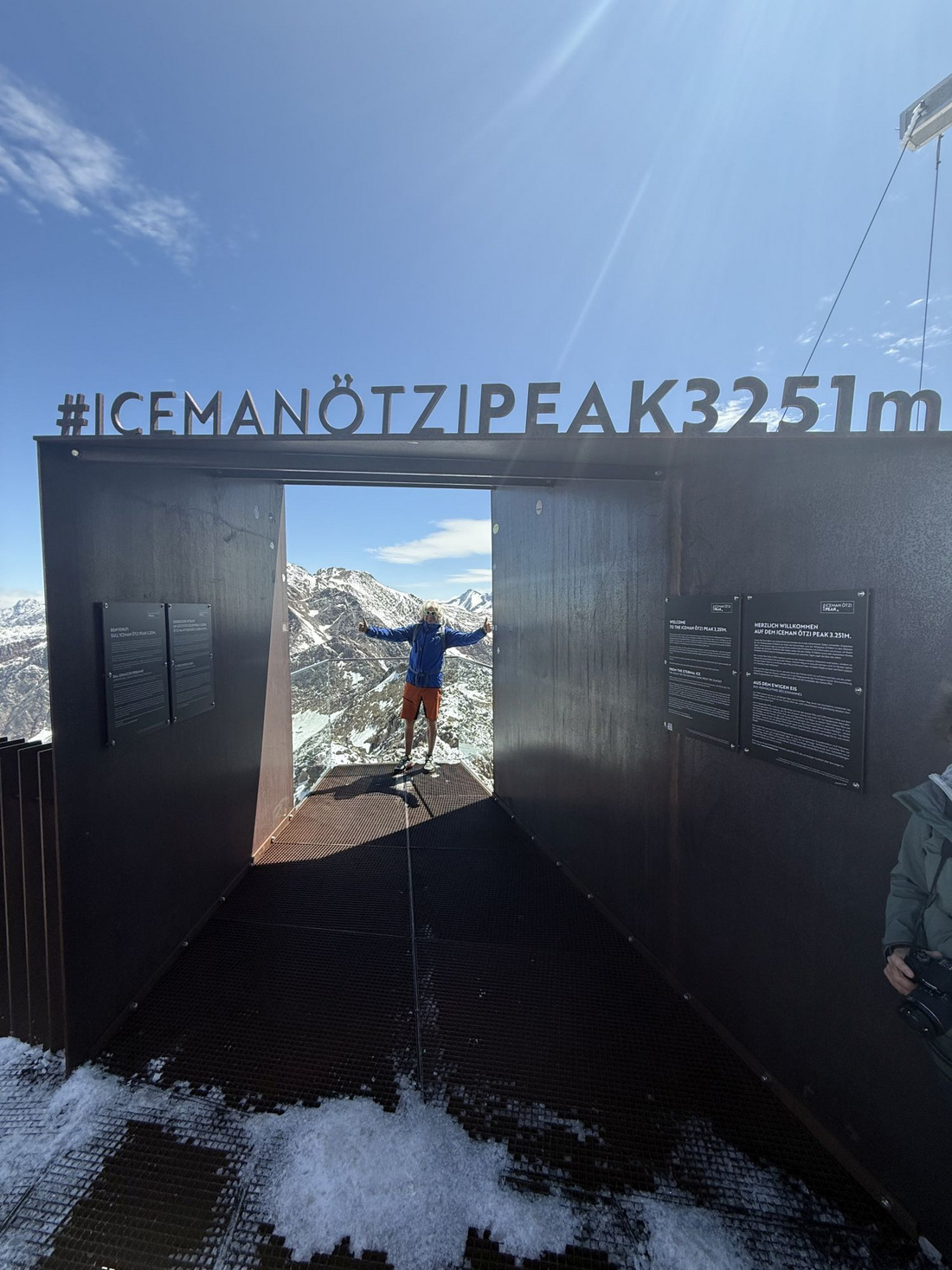 Punkt widokowy Iceman Ötzi Peak 3251m urysta z uniesionymi rękami stoi na metalowej platformie widokowej wewnątrz dużej stalowej konstrukcji z napisem Iceman Ötzi Peak 3251m, w tle widać ośnieżone góry.