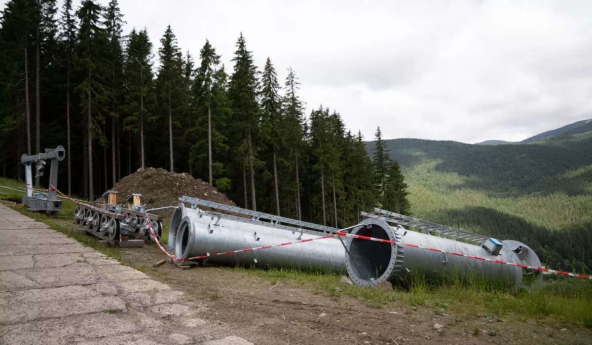Elementy słupa nowej kolei linowej w Szpindlerowym Młynie Metalowe rury stanowiące trzon słupa kolei linowej leżące na trawie przy betonowej drodze. Elementy są zabezpieczone biało-czerwoną taśmą ostrzegawczą.