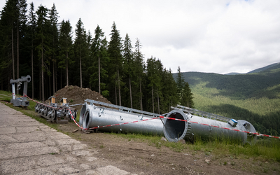 Metalowe rury stanowiące trzon słupa kolei linowej leżące na trawie przy betonowej drodze. Elementy są zabezpieczone biało-czerwoną taśmą ostrzegawczą.