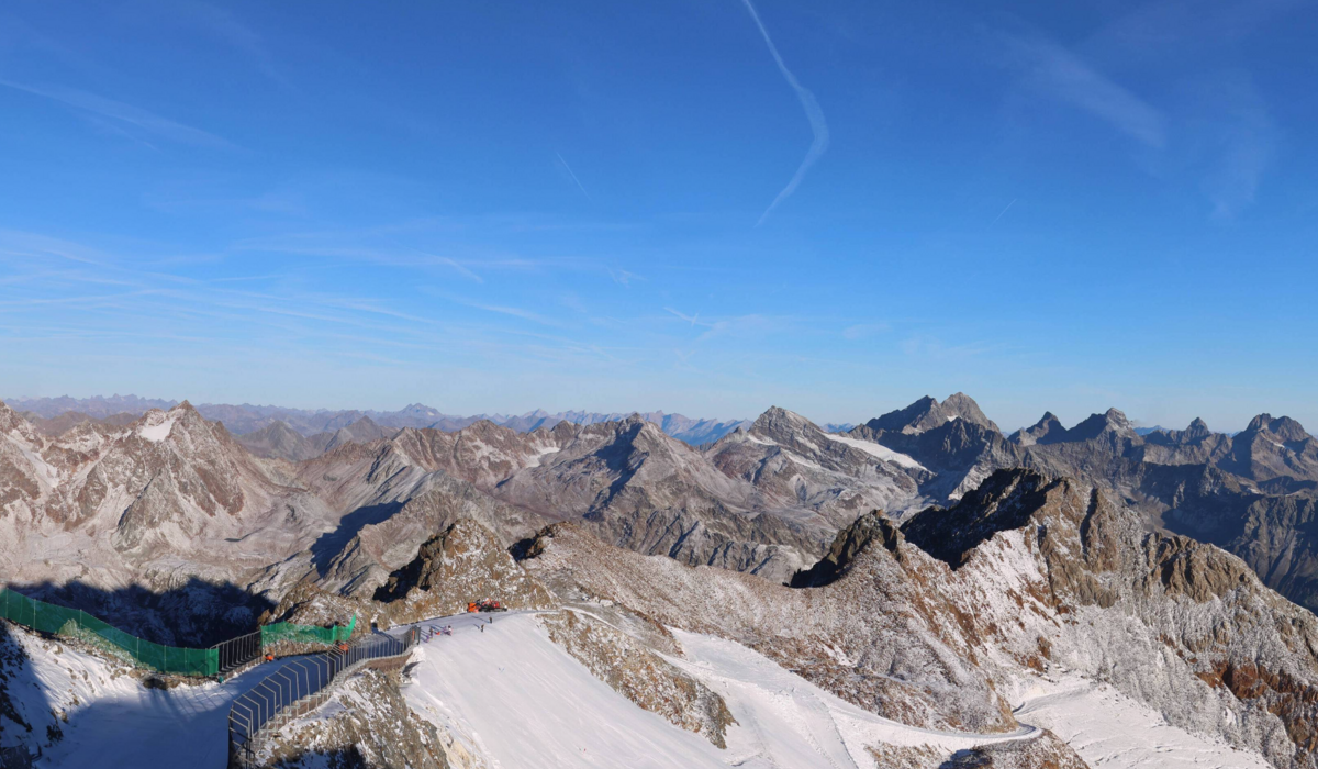 Obraz z kamery internetowej z Café 3440 na lodowiec Pitztal (25 09 2026 g. 8:30/ źródło https://www.pitztaler-gletscher.at/en/webcam.html#webcam Szczyty górskie na tle niebieskiego nieba, w górach przygotowane trasy narciarskie na lodowcu