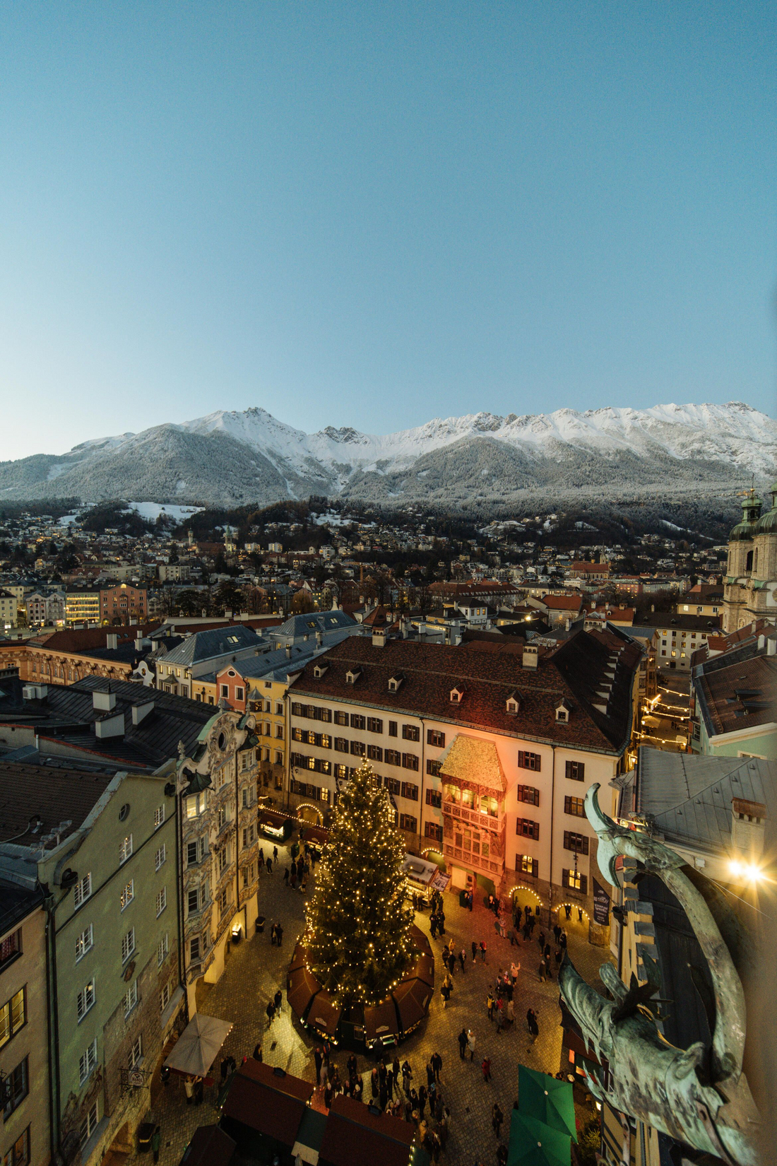 Fot.: Innsbruck Tourismus / Martin Venier Widok z góry na jarmark bożonarodzeniowy w Innsbrucku z choinką i ośnieżonymi Alpami w tle o zmierzchu.