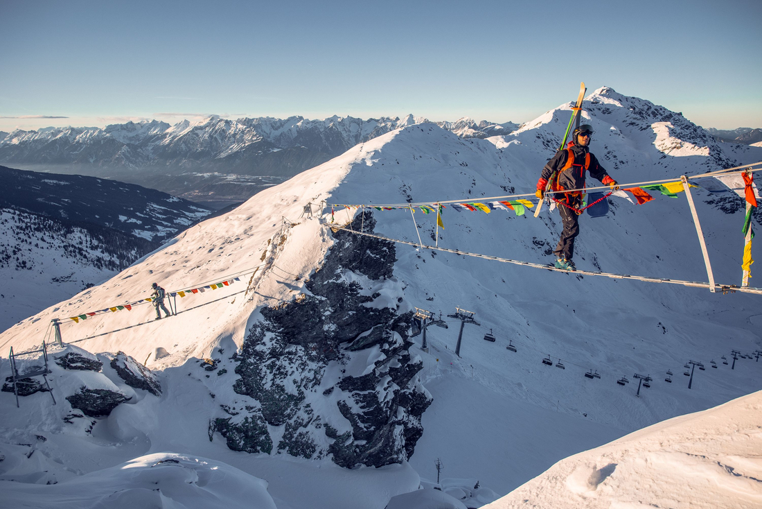 Ekstremalne przejście przez most linowy „Skywalk” na lodowcu Stubai, z wspinaczem niosącym narty i panoramą Alp w tle. Fot. TVB Stubai Tirol / Christian Eberl Wspinacz w kasku i z nartami przypiętymi do plecaka ostrożnie przechodzi po wiszącym moście linowym rozpiętym między skalistymi szczytami. Most ozdobiony jest kolorowymi flagami modlitewnymi. W tle widać rozległą panoramę ośnieżonych gór pod czystym niebem.