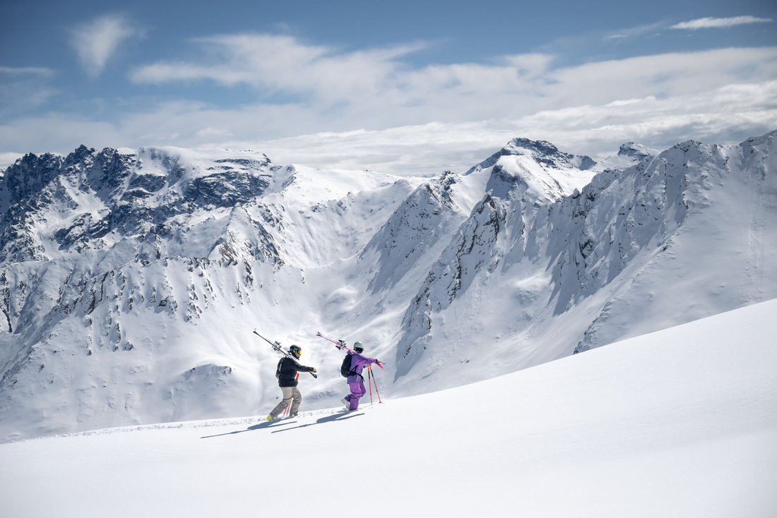 (c) TVB Paznaun - Ischgl Dwóch narciarzy z nartami na plecach wchodzi po naśnieżonym zboczu w stronę grani, z panoramą gór Paznaun w tle.