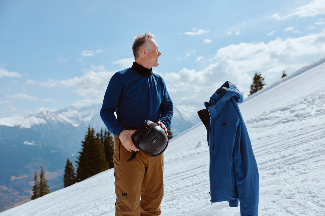  Mężczyzna w niebieskiej bluzie i brązowych spodniach trzyma kask na ośnieżonym stoku; obok znajduje się niebieska kurtka Helly Hansen.