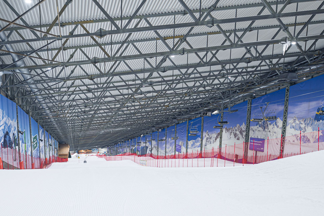 Główna trasa narciarska w hali Snow Arena Druskienniki Szeroka, idealnie przygotowana trasa narciarska pokryta technicznym śniegiem wewnątrz hali z widocznym potężnym stalowym zadaszeniem i barierami ochronnymi.