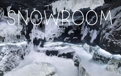 Wnętrze lodowej jaskini lub pomieszczenia typu snowroom z czarnymi skałami pokrytymi grubą warstwą śniegu i wiszącymi soplami lodu. Na górze obrazu widnieje duży, biały napis „SNOWROOM”.