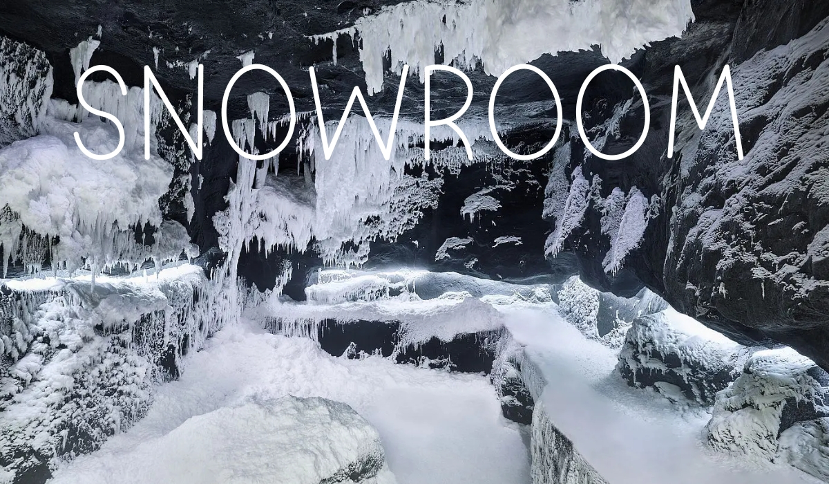 Snowroom by Technoalpin Wnętrze lodowej jaskini lub pomieszczenia typu snowroom z czarnymi skałami pokrytymi grubą warstwą śniegu i wiszącymi soplami lodu. Na górze obrazu widnieje duży, biały napis „SNOWROOM”.