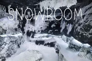 Wnętrze lodowej jaskini lub pomieszczenia typu snowroom z czarnymi skałami pokrytymi grubą warstwą śniegu i wiszącymi soplami lodu. Na górze obrazu widnieje duży, biały napis „SNOWROOM”.