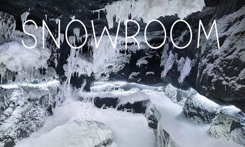 Snowroom by Technoalpin Wnętrze lodowej jaskini lub pomieszczenia typu snowroom z czarnymi skałami pokrytymi grubą warstwą śniegu i wiszącymi soplami lodu. Na górze obrazu widnieje duży, biały napis „SNOWROOM”.