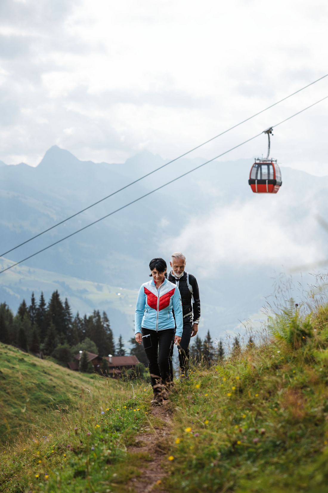 Kitzbühel, Hahnenkammbahn (c)Tirol Werbung, Roman Huber Para podchodząca górską ścieżką nad nimi czerwona gondola kolei linowej