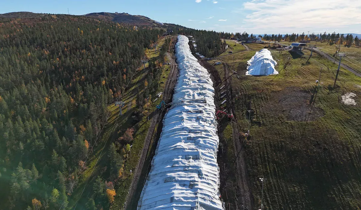 Przygotowywanie Trasy Narciarskiej Metodą Snow Farming (Przechowywanie Śniegu) Widok z lotu ptaka na zielone zbocze górskie jesienią, na którym usypano długi pas magazynowanego śniegu pod białą płachtą ochronną, z pracującymi koparkami.
