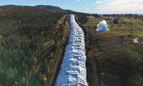 Przygotowywanie Trasy Narciarskiej Metodą Snow Farming (Przechowywanie Śniegu) Widok z lotu ptaka na zielone zbocze górskie jesienią, na którym usypano długi pas magazynowanego śniegu pod białą płachtą ochronną, z pracującymi koparkami.