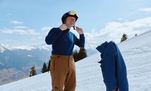 Przygotowanie do jazdy – Helly Hansen na stoku Narciarz w niebieskiej bluzie i brązowych spodniach zapina pasek kasku na stoku, obok niebieskiej kurtki Helly Hansen.