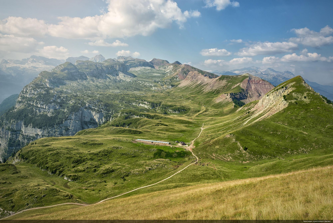 Rozległa panorama zielonych górskich grzbietów z wijącą się ścieżką i schroniskiem pośrodku doliny.