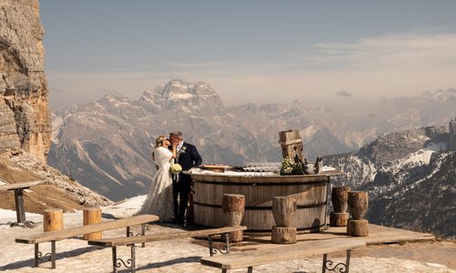 Narty i ślub podczas jennych wakacji :) / cortina.dolomiti.org Szerokie ujęcie pary młodej całującej się na tarasie widokowym w Alpach. Panna młoda ma na sobie białą suknię i futrzaną etolę, pan młody ciemny garnitur. Stoją obok drewnianego baru w kształcie beczki, a w tle rozpościera się spektakularny widok na surowe, ośnieżone pasmo górskie Dolomitów pod jasnym niebem.