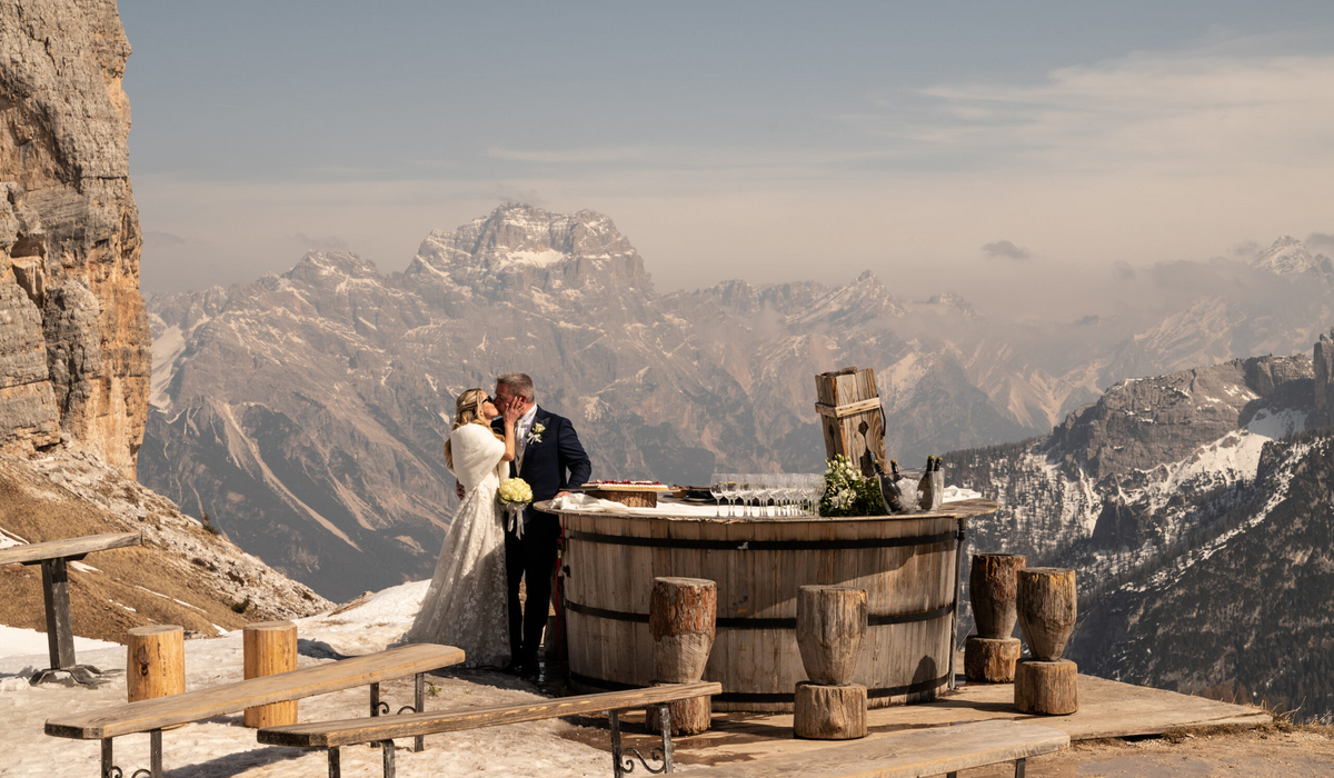 Narty i ślub podczas jennych wakacji :) / cortina.dolomiti.org Szerokie ujęcie pary młodej całującej się na tarasie widokowym w Alpach. Panna młoda ma na sobie białą suknię i futrzaną etolę, pan młody ciemny garnitur. Stoją obok drewnianego baru w kształcie beczki, a w tle rozpościera się spektakularny widok na surowe, ośnieżone pasmo górskie Dolomitów pod jasnym niebem.