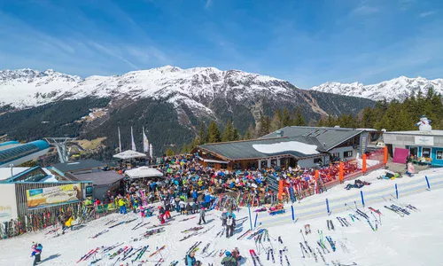 Wiosenne party na stoku w Kappl © TVB Paznaun – Ischgl Tłum narciarzy przy górskiej restauracji na stoku narciarskim, z widokiem na ośnieżone szczyty i wyciąg narciarski.