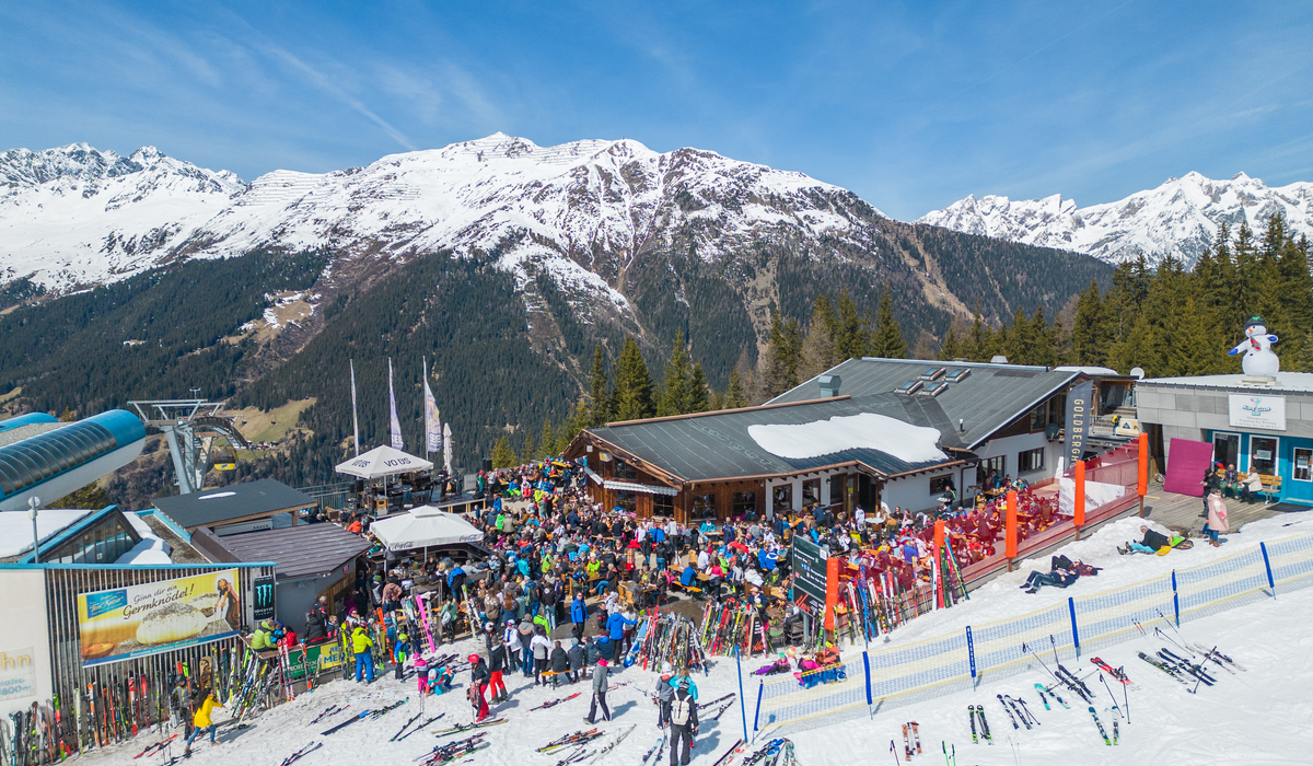 Wiosenne party na stoku w Kappl © TVB Paznaun – Ischgl Tłum narciarzy przy górskiej restauracji na stoku narciarskim, z widokiem na ośnieżone szczyty i wyciąg narciarski.