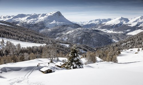 Malownicza zimowa sceneria w austriackich Alpach ze śladami na śniegu w pobliżu tradycyjnych górskich chat. Fot. (c)TVB Rudi Wyhlidal Zimowa panorama górskiej doliny pokrytej śniegiem. W pierwszym planie widać kilka małych, drewnianych chat i ośnieżone choinki. Na zboczach rozciąga się gęsty las iglasty, a w tle wznoszą się majestatyczne, skaliste szczyty.