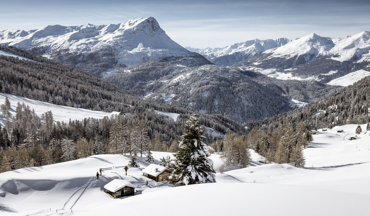 Malownicza zimowa sceneria w austriackich Alpach ze śladami na śniegu w pobliżu tradycyjnych górskich chat. Fot. (c)TVB Rudi Wyhlidal Zimowa panorama górskiej doliny pokrytej śniegiem. W pierwszym planie widać kilka małych, drewnianych chat i ośnieżone choinki. Na zboczach rozciąga się gęsty las iglasty, a w tle wznoszą się majestatyczne, skaliste szczyty.