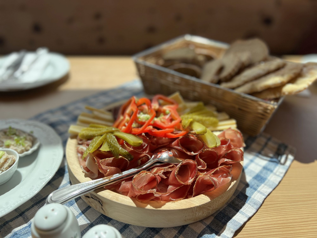 Tradycyjna deska wędlin i serów w górskim schronisku Drewniany talerz pełen zwiniętych plastrów szynki dojrzewającej, udekorowany czerwoną papryką i ogórkami kiszonymi, postawiony na obrusie w niebieską kratę.